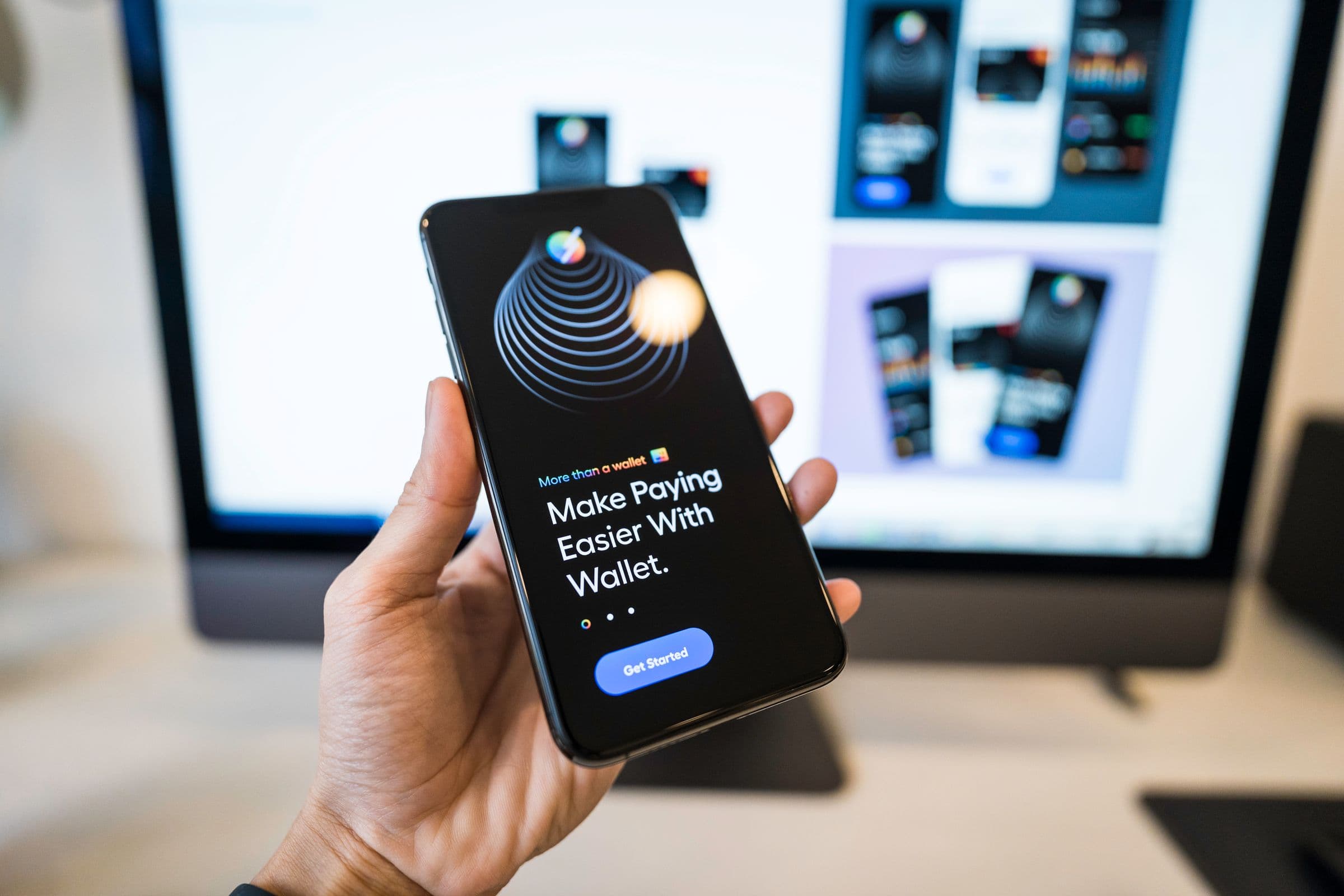 Elinde uygulama ekranı açık telefon; arkadaki monitörde aynı uygulamanın tasarım ekranları